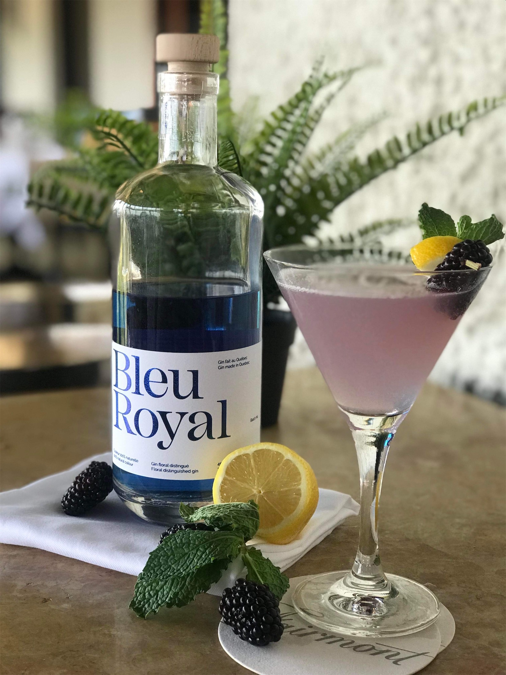 a cocktail with blackberry and lemon garnish with a bottle of bleu royal next to it